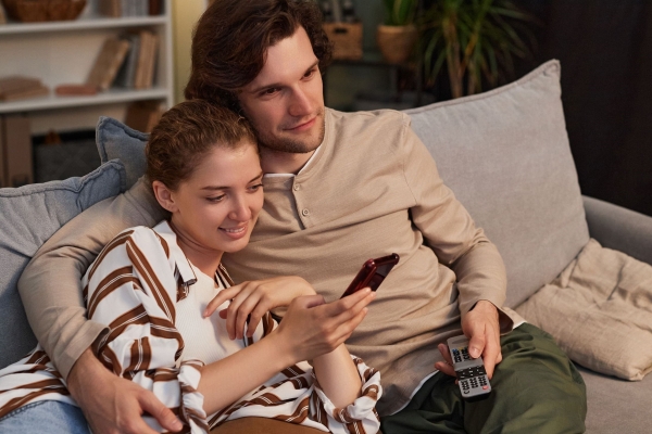 A white man with short dark hair wearing a light top and dark trousers holding a TV remote control sits on a sofa with his arm round a white woman who is wearing a striped top and light jeans and is looking at a mobile phone